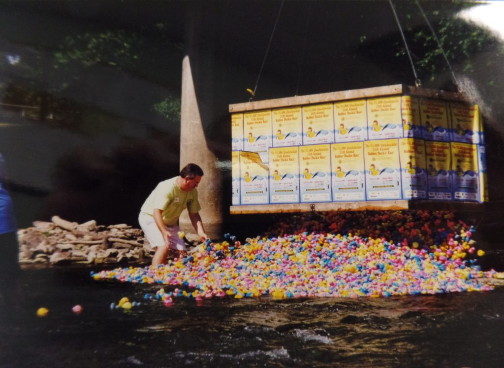 Image shows a man in the river surrounded by small rubber ducks that have just been dumped in by a giant container