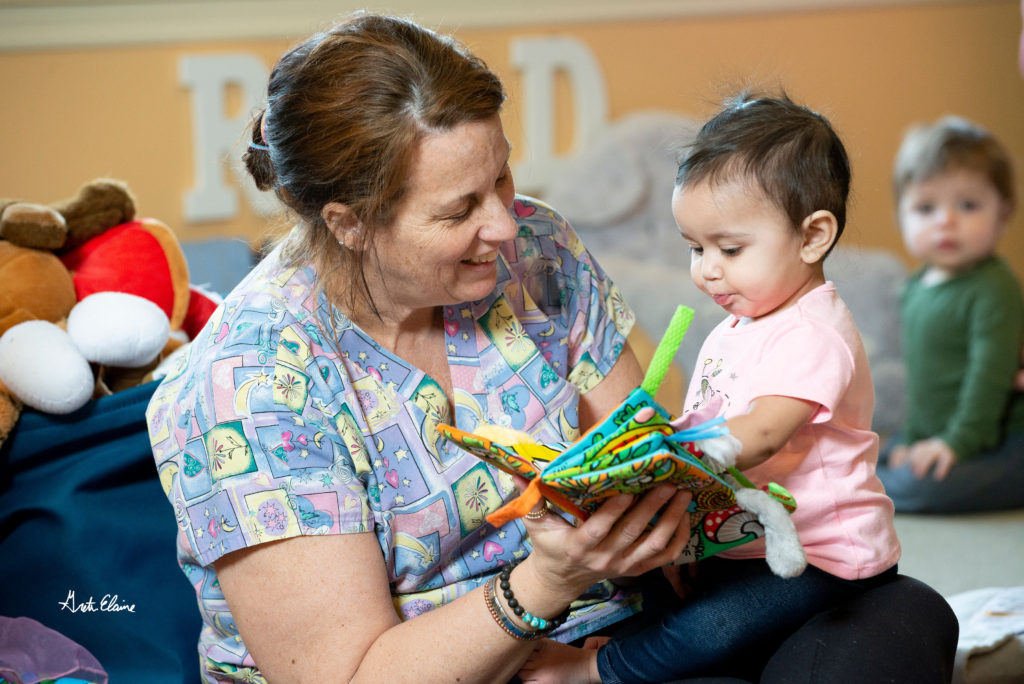 Image shows an infant being read to by a staff member in the child care center