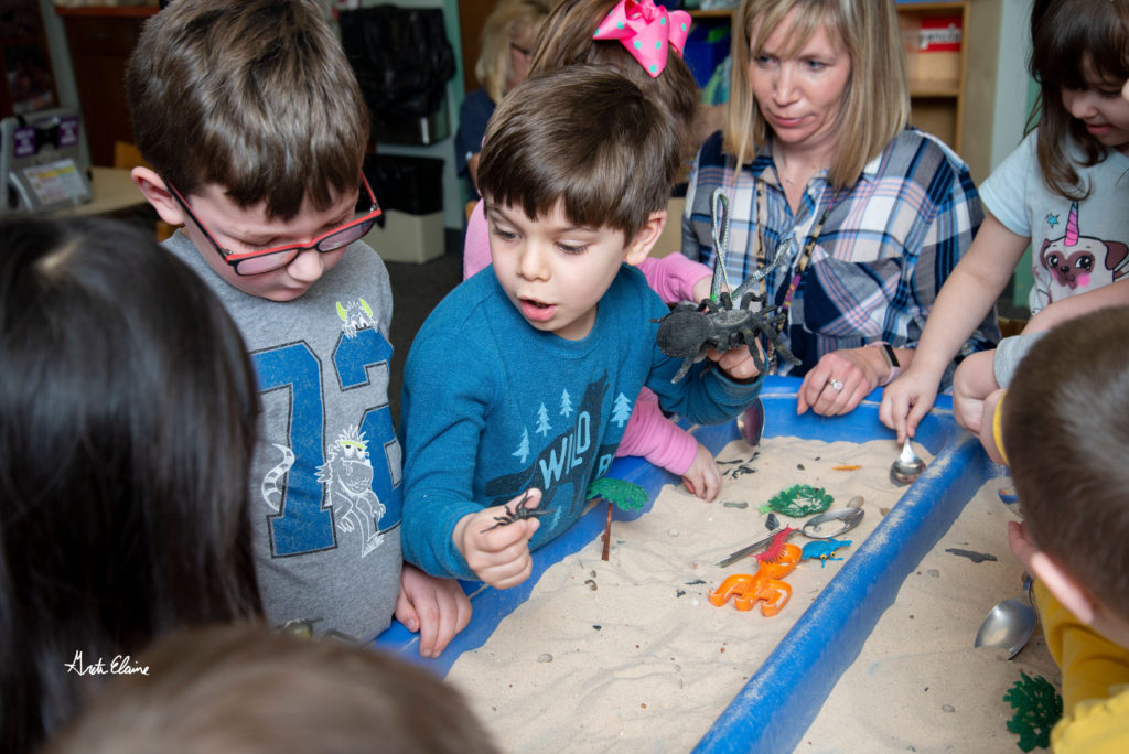 Several young children gather around an indoor sandbox filled with toys playing together while a woman caregiver supervises. 