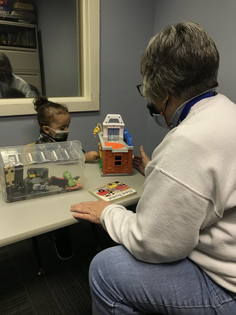 Image shows a young girl and her therapists working through play with toy cars, a toy car wash station, and a book called "I love trucks!"