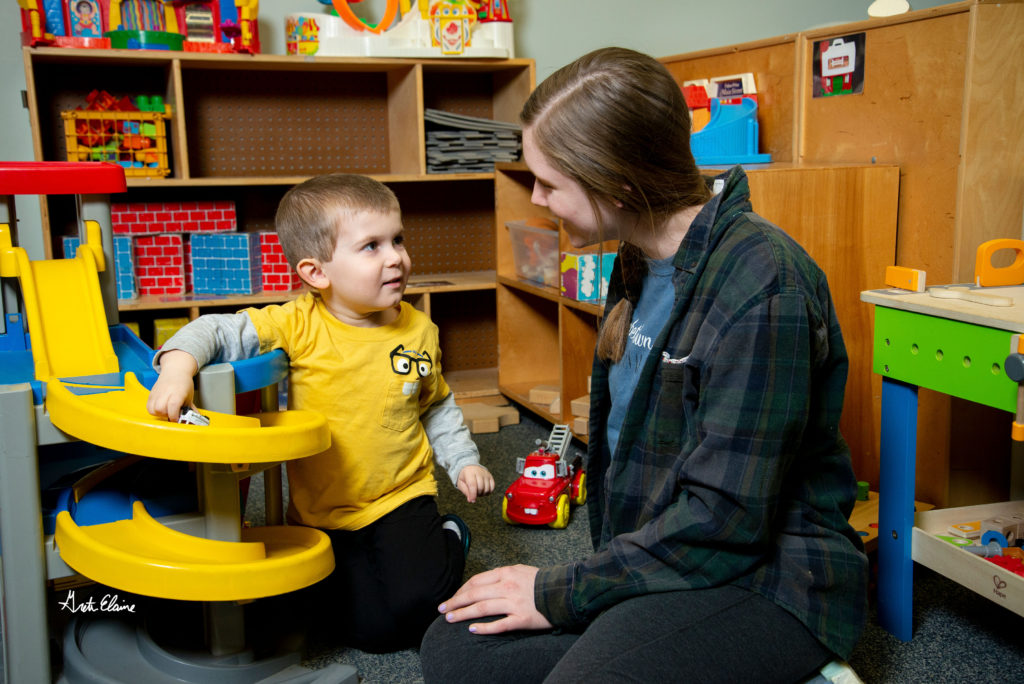 Preschool aged boy and young woman sit on the floor together while the young boy plays with a toy car on a ramp