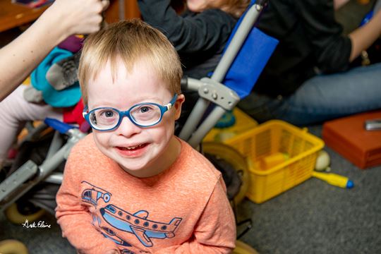 Young boy wearing glasses and sitting on the floor smiles up at the camera 