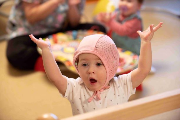 Infant girl with cochlear implants and a pink bonnet raising her hands in excitement while infant boy and staff member play together in the background