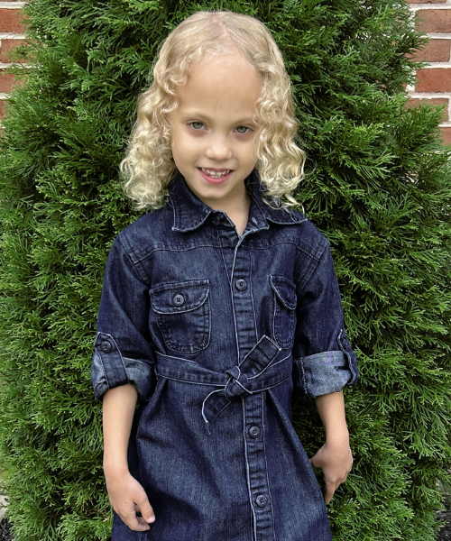 Avery smiling standing in front of a small pine tree wearing a denim dress.
