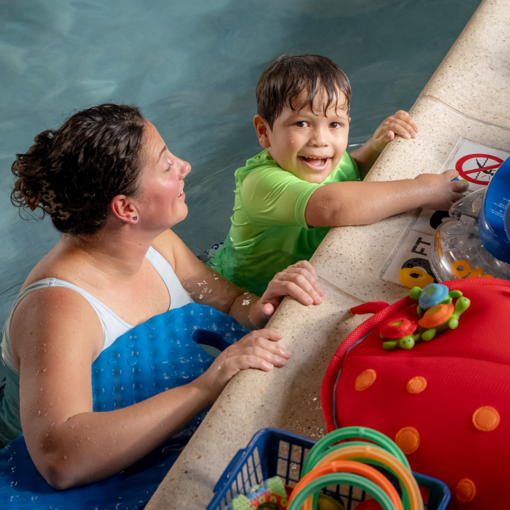 young boy in a pool, at the edge, smiling up at the camera next to his female therapist