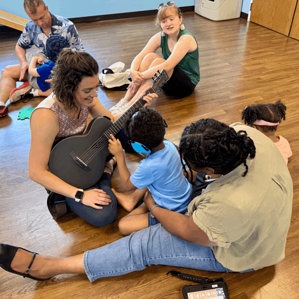 Little boy sitting in moms lap plays guitar with instructor in music therapy camp