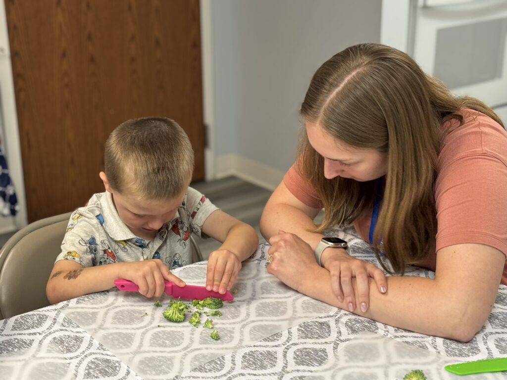 little boy at a table cutting broccoli with camp counselor