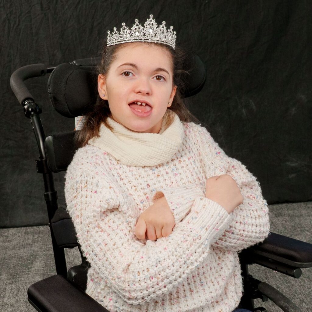 young girl in a white sweater and diamond tiara sitting in wheelchair in front of black backdrop