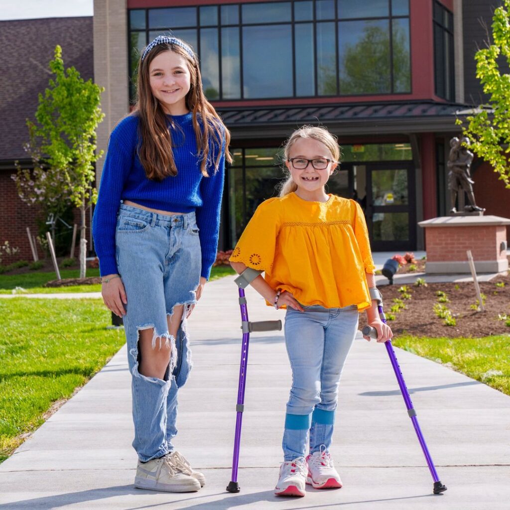two girls standing in front of Schreiber building. One with crutches.