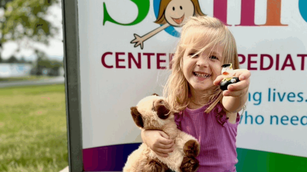 A child sitting and smiling in front of a Schreiber Center of Pediatric Development sign holding a stuffed animial.
