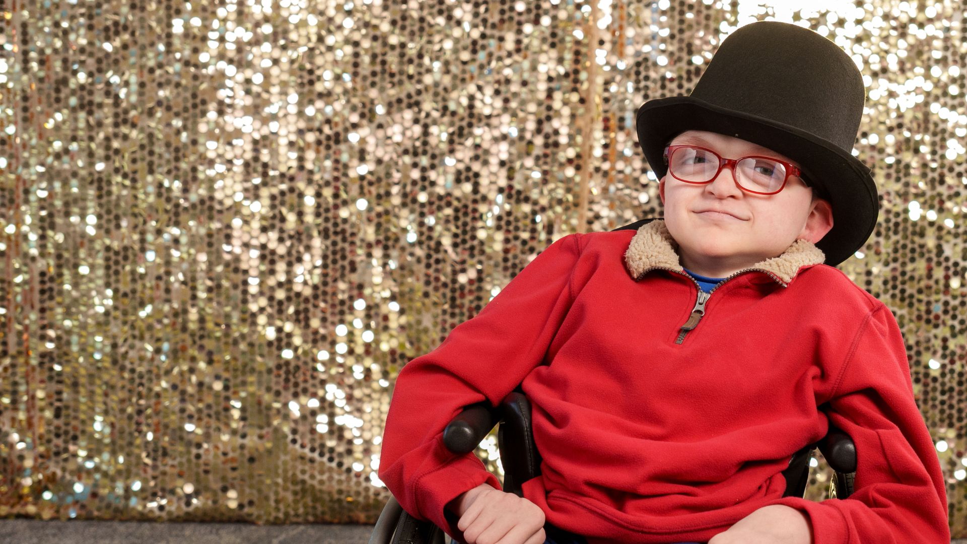 young boy in red sweater and black top hat sitting in wheelchair in front of sparkly gold backdrop