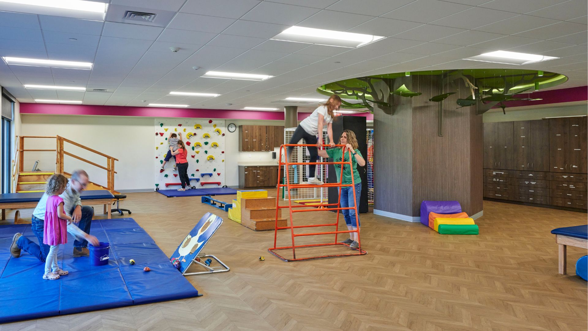 young girl climbing an orange ladder with the help of her physical therapist while a girl plays a bean bag game and a boy climbs the rock wall in the backround