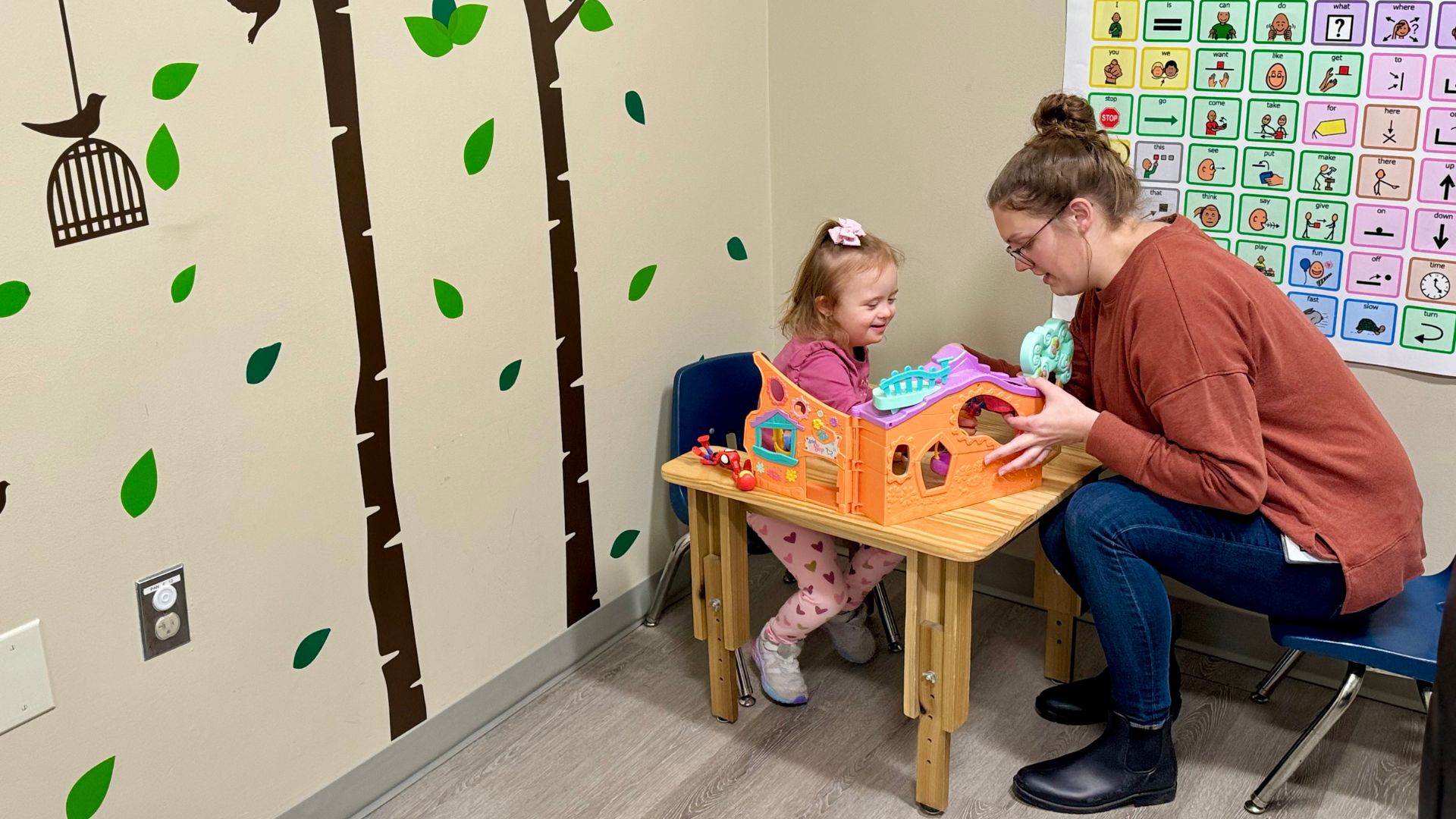 A child with a bow in her hair working at a table with her speech therapist.