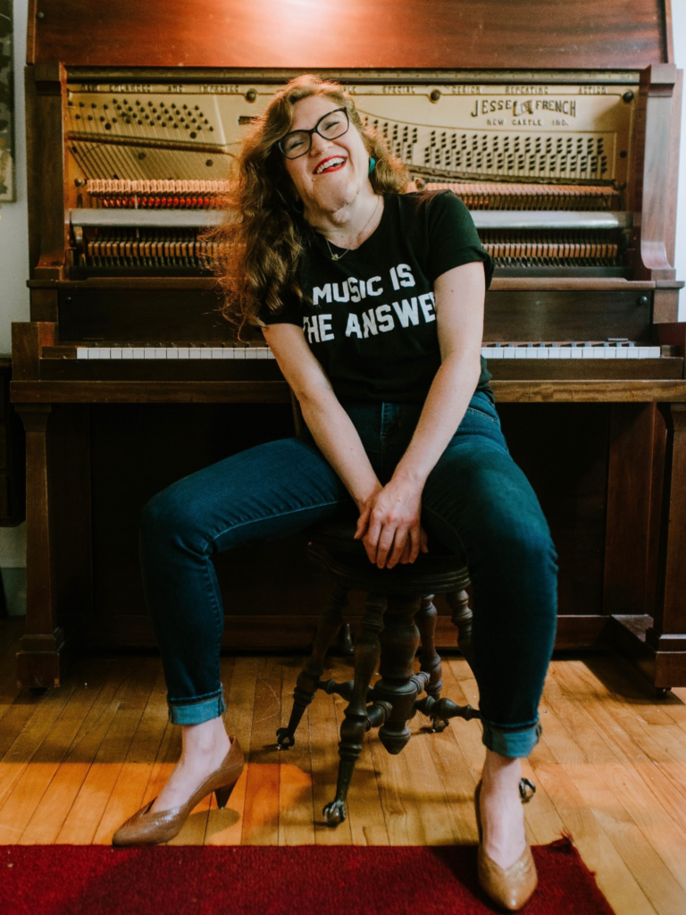 Woman sitting in front of a piano wearing a shirt that says "Music is the Answer"
