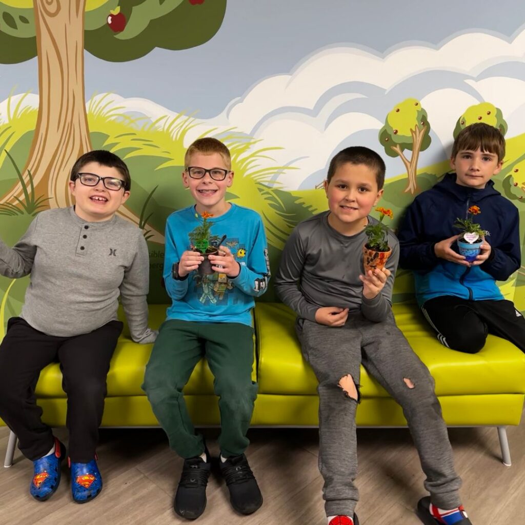 Four young boys sitting on a bench in front of a mural holding up their potted plants for mothers day